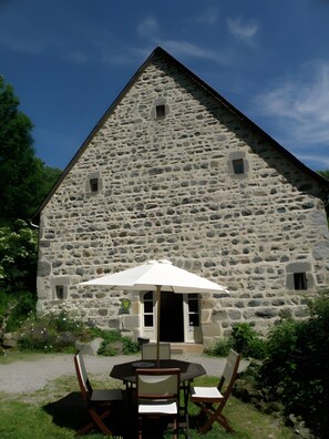 A stone building with a triangular roof, a small window, and a door. In front, there is a round table with chairs and a white umbrella. - Holiday Home 'Chambres D'hôtes Et Gîte Du Domaine' with Mountain View and Wi-Fi (Bagnols)