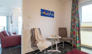 Ein Zimmer mit einem roten Sofa, einem kleinen Tisch, zwei Stühlen, einem Bild an der Wand und einem Fenster mit Vorhängen.