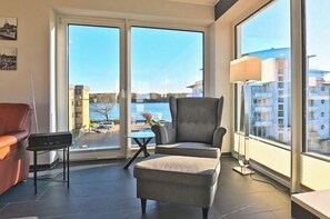 A modern living room with a grey armchair, a small table, and a sofa. Large windows offer a view of buildings and a body of water. - Holiday apartment with 2 bedrooms (Heiligenhafen)