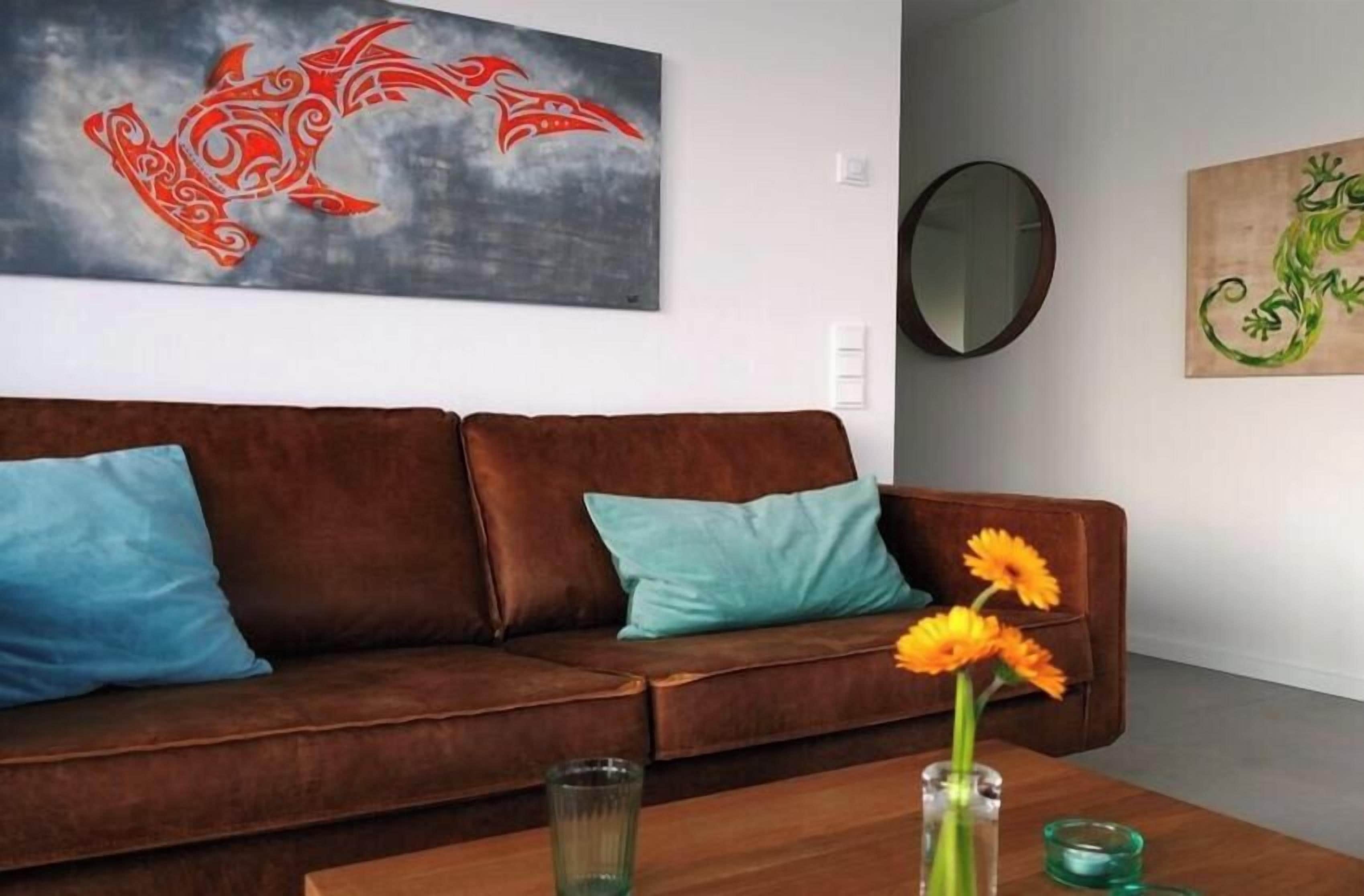 A brown leather sofa with teal cushions, a glass vase with flowers, and two paintings on the wall.