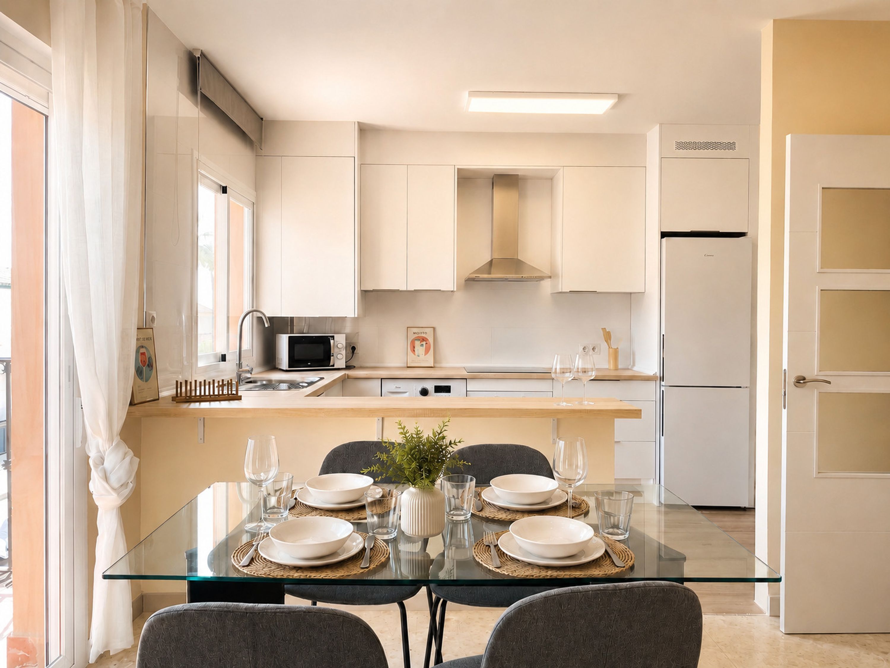Una cocina moderna con gabinetes blancos, un microondas y un refrigerador. Hay una mesa de comedor con platos, vasos y un florero.
