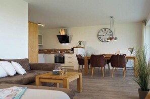 A modern living room with a sofa, coffee table, and dining area with chairs and a table. A clock is on the wall, and there is a plant in the corner.