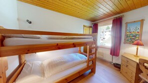 A bunk bed room with a wooden ceiling, a window with curtains, a painting on the wall, and a small wooden dresser. - Apartment house on the hillside of Celerina. (Celerina/Schlarigna)