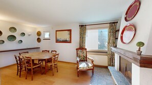 A dining area with a round wooden table and chairs, a fireplace, and decorative plates on the wall. - Apartment house on the hillside of Celerina. (Celerina/Schlarigna)
