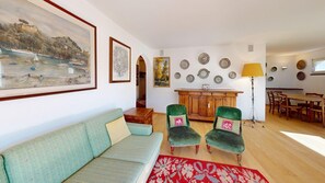 A living room with a sofa, two armchairs, a dining table with chairs, a lamp, and various wall decorations including framed pictures and plates. - Apartment house on the hillside of Celerina. (Celerina/Schlarigna)