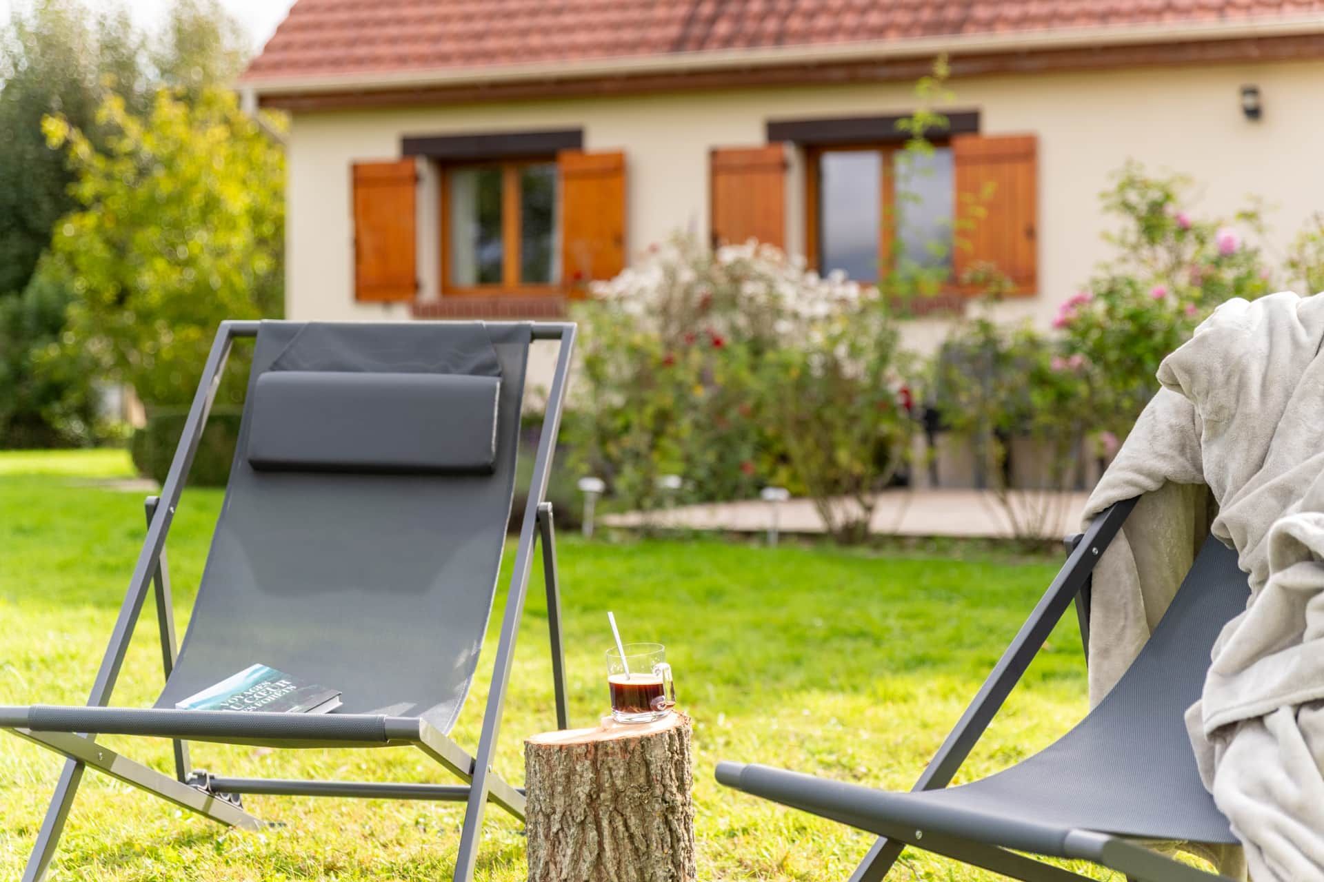 Un fauteuil avec un coussin et un verre posé sur une souche d’arbre, installé dans un espace herbeux avec une maison en arrière-plan.