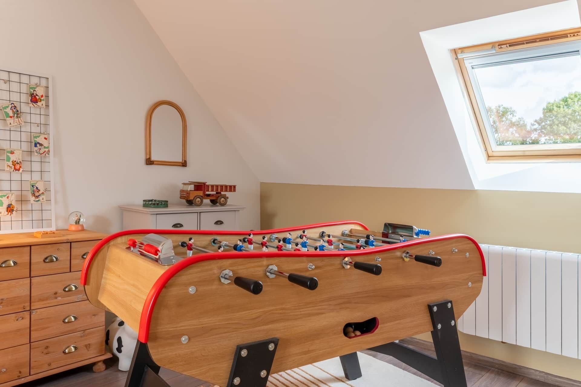 Une table de baby-foot dans une pièce dotée d’un plafond en pente, d’une fenêtre offrant une vue dégagée et d’une grille murale où sont accrochées des photos.