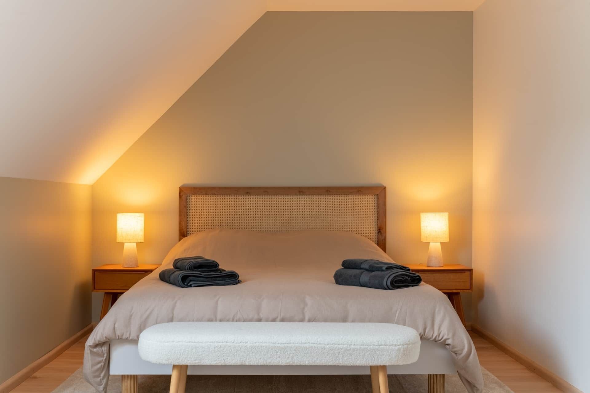 Ein Schlafzimmer mit schrägverlaufender Decke, einem Bett mit Kopfteil, zwei Nachttischlampen, einer Bank und gefalteten Handtüchern auf dem Bett.
