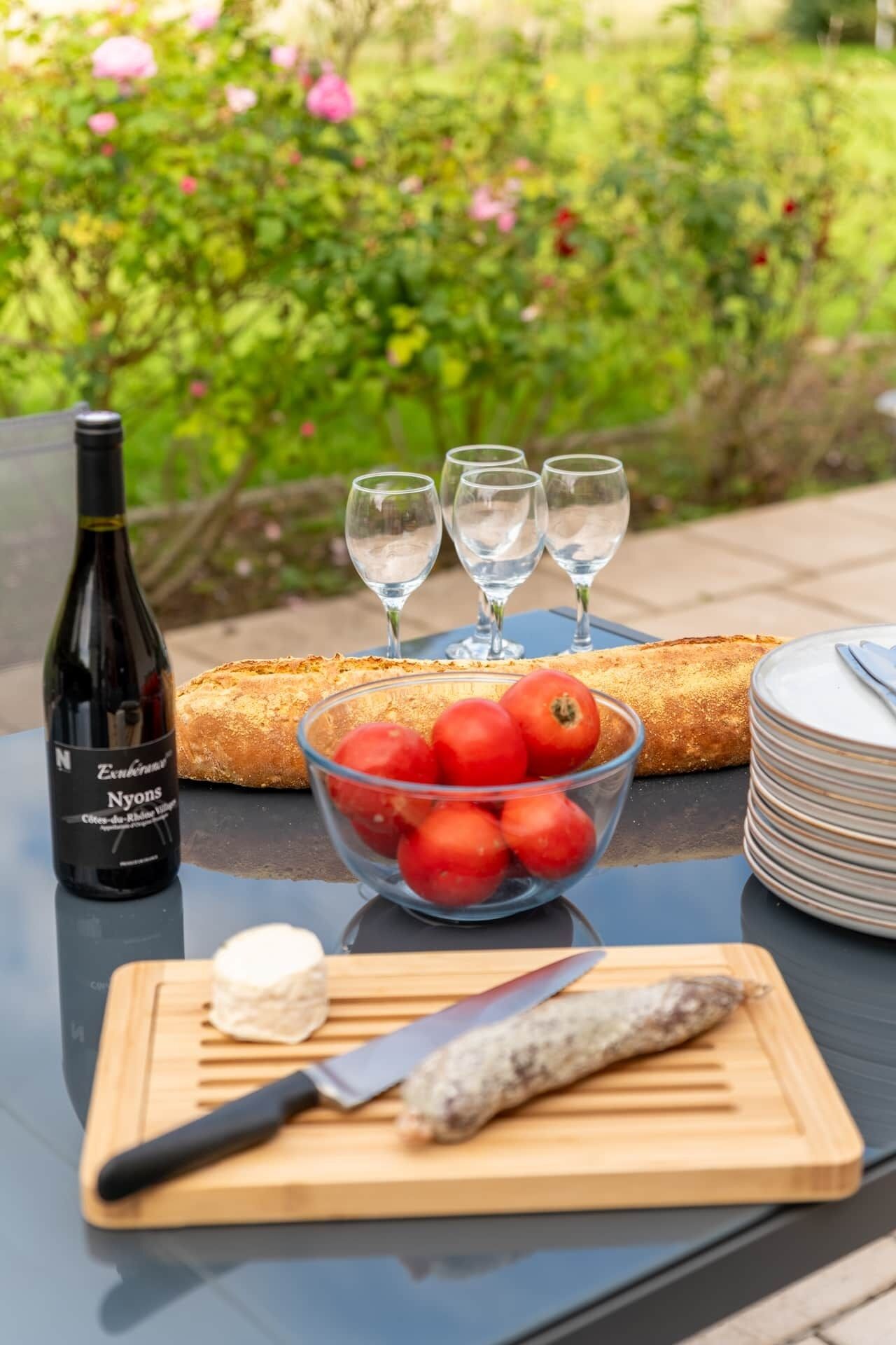 A table with a bottle of wine, glasses, a bowl of tomatoes, a baguette, plates, a cutting board with a knife, and a piece of fish.