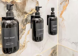 Trois flacons noirs contenant du gel douche et du shampoing, des produits essentiels pour l’hygiène corporelle, sont posés sur un mur en marbre.