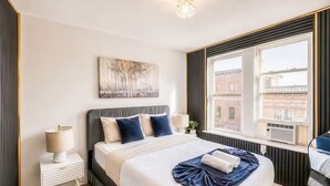 Habitación de hotel con una cama grande, una mesita de noche, una lámpara, un ventanal con vistas a edificios y una obra de arte colgada en la pared.