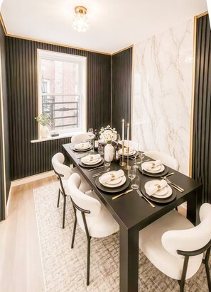 Un espace repas avec une longue table dressée pour quatre personnes, des murs sombres, une fenêtre offrant une vue et un mur d’accent en marbre.