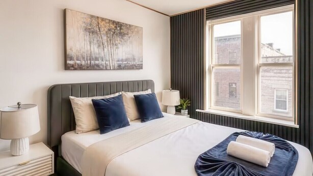 Habitación de hotel con cama, lámparas de noche, una ventana con vistas y un cuadro en la pared.