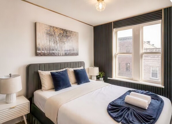 Habitación de hotel con cama, lámparas de noche, una ventana con vistas y un cuadro en la pared.