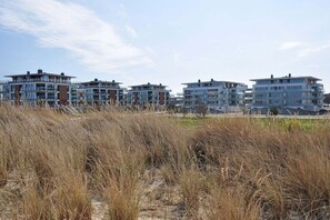 A row of modern multi-story buildings with a flat roof design, situated on a sandy terrain with dry grass in the foreground. - Holiday apartment with 2 bedrooms (Heiligenhafen)