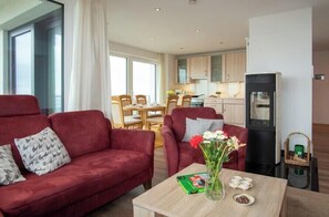 Ein Wohnzimmer mit einem roten Sofa, einem Holztisch und einer Vase mit Blumen. Im Hintergrund ist eine Küche mit Holzschränken und einem Kühlschrank zu sehen.