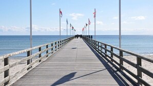 Un muelle de madera que se adentra en el mar, con banderas a ambos lados, que conduce a una playa.