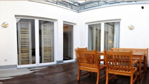 Un espacio al aire libre cubierto con una mesa y sillas de madera, barandilla de cristal y pared blanca con ventanas.