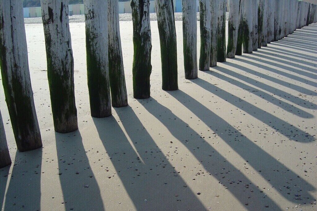 Eine Reihe vertikaler Holzbalken wirft Schatten auf eine sandige Fläche.