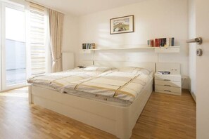 A bedroom with a bed, bedside tables, a window with curtains, and a wall-mounted shelf with books. - Holiday apartment in Norderney with Wi-Fi (Norderney)