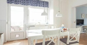 A bright room with a white table and chairs, a window with curtains, and a hanging light fixture. - Mariechen am Meer Apt. 1 - Norddeich (Norddeich)