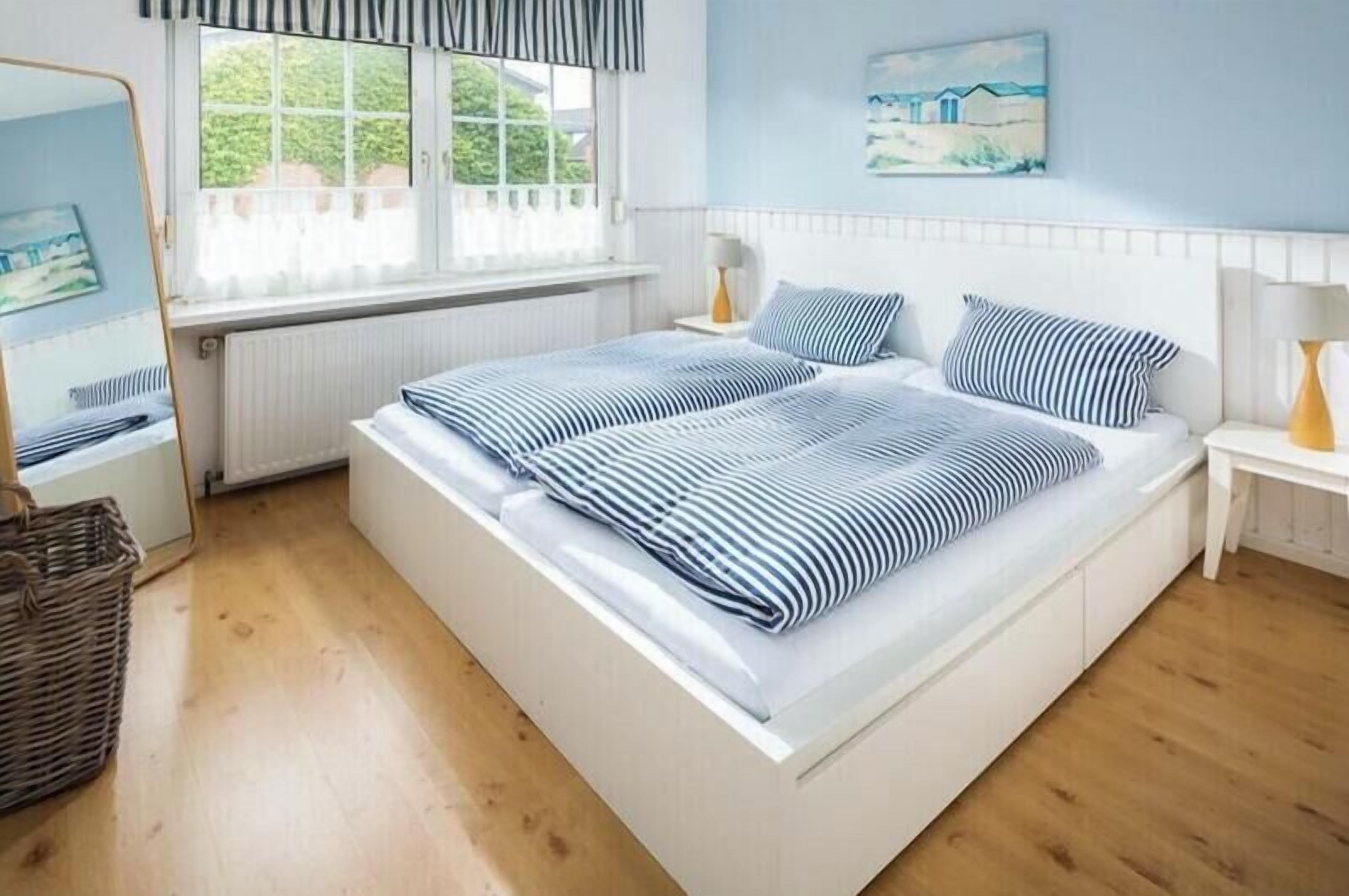 A double bed with striped bedding, a bedside table with a lamp, a window with curtains, and a painting on the wall.