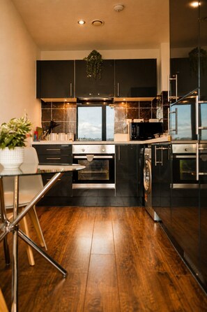 A modern kitchen with dark cabinetry, a stainless steel oven, a microwave, and a glass dining table with a plant. - Spacious Liverpool City Centre Apartment with View Managed by Sidemersey Livings (Liverpool)