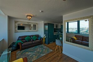 Ein Wohnzimmer mit braunem Ledersofa, einem grün gemusterten Teppich, einem Essbereich mit Tisch und Stühlen, einer Küche mit türkisfarbenen Schränken und einem großen Fenster mit Blick auf die Stadt.