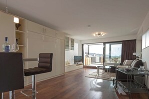 Ein modernes Wohnzimmer mit einer Couch, einem Couchtisch und einem Fernseher. Ein Balkon bietet Blick auf die Stadt.