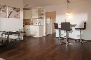 Una cocina moderna con gabinetes blancos, un comedor con un carrito metálico y dos taburetes de bar, y una lámpara colgante.