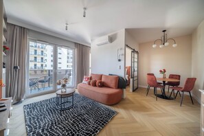 A modern living room with a sofa, a dining table with chairs, a patterned rug, and a view of a building through large windows. - Kierbedzia Comfort Stay, Mokotów, Warsaw (Warszawa)