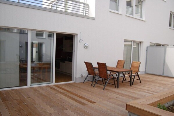 A patio with a wooden table and chairs, a sliding glass door leading to a kitchen, and a white building with windows. - Holiday apartment in Norderney with Wi-Fi (Norderney)