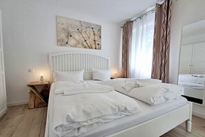 Een slaapkamer met een wit bed, een nachtkastje met een lamp, een wanddecoratie en een raam met gordijnen.