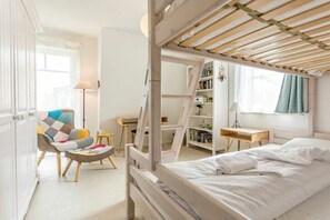 Een slaapkamer met een stapelbed, een stoel met een kleurrijk kussen, een nachtkastje, een boekenkast en een raam met gordijnen.