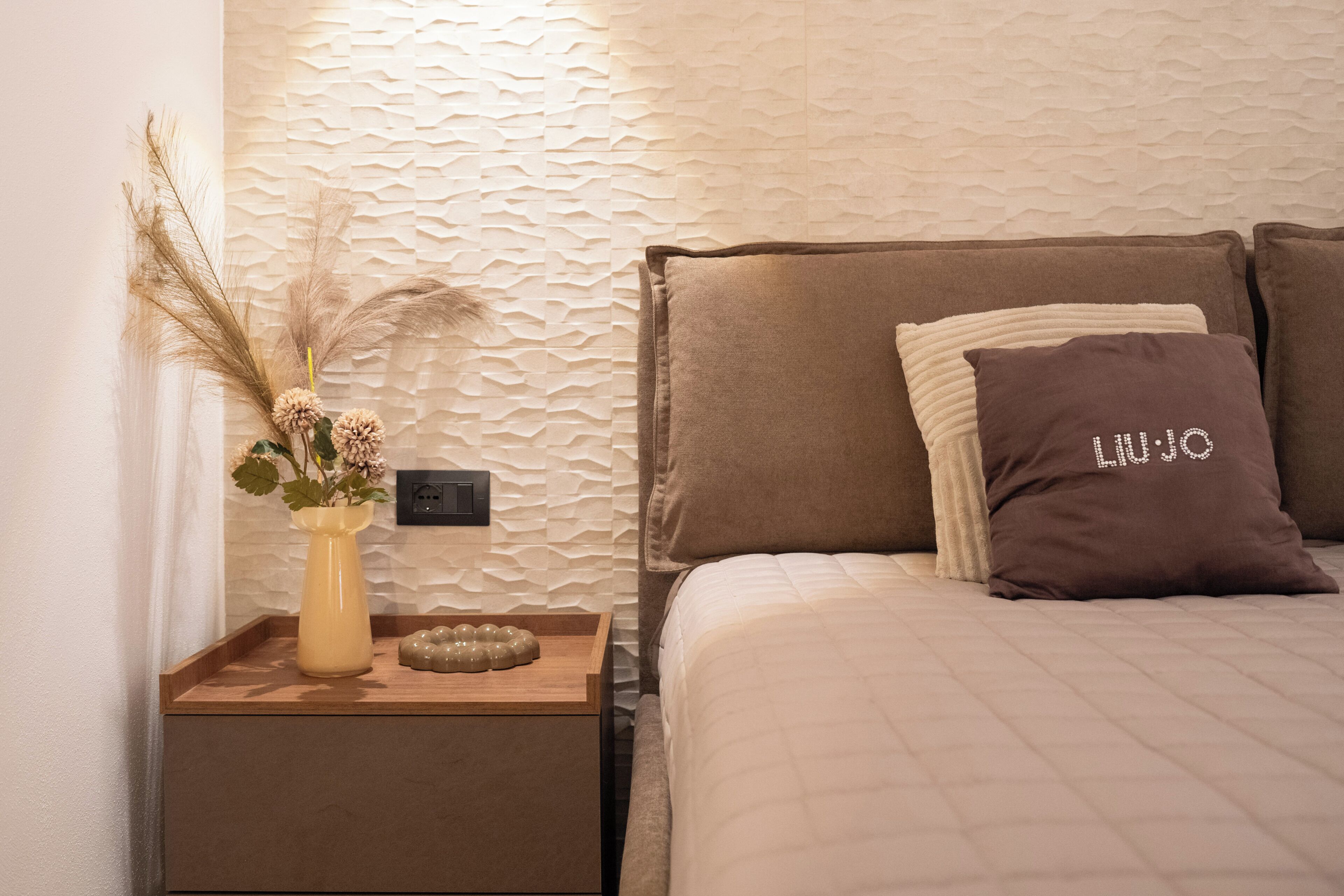 Un dormitorio ordenado con cama, almohadas, una mesita de noche, un florero con plantas secas y una pared con textura.