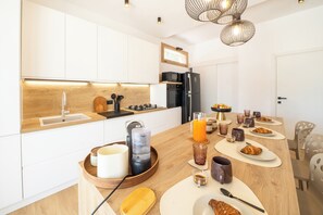Una cocina moderna con armarios blancos, encimera de madera y comedor equipado con platos, vasos y una máquina de café.