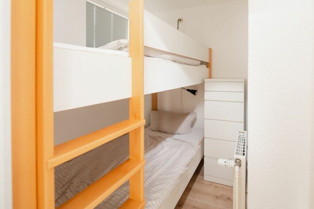 Habitación con literas, una escalera de madera, un radiador blanco y una ventana con persianas.