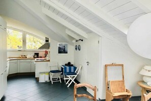 Een compacte keuken met witte kasten, een eethoek met een blauw-wit geruit tafelkleed en een houten stoel.
