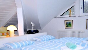 Quarto com teto inclinado, cama com roupa de cama listrada, abajur, quadro na parede e radiador.