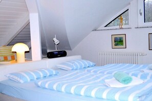 Een slaapkamer met een schuine kap, een bed met gestreepte lakens, een bedlampje, een ingelijst beeld aan de muur en een radiator.