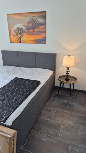 Ein Schlafzimmer mit einem Bett, einem Nachttisch mit Lampe, einem Wandbild und einem gefliesten Boden.
