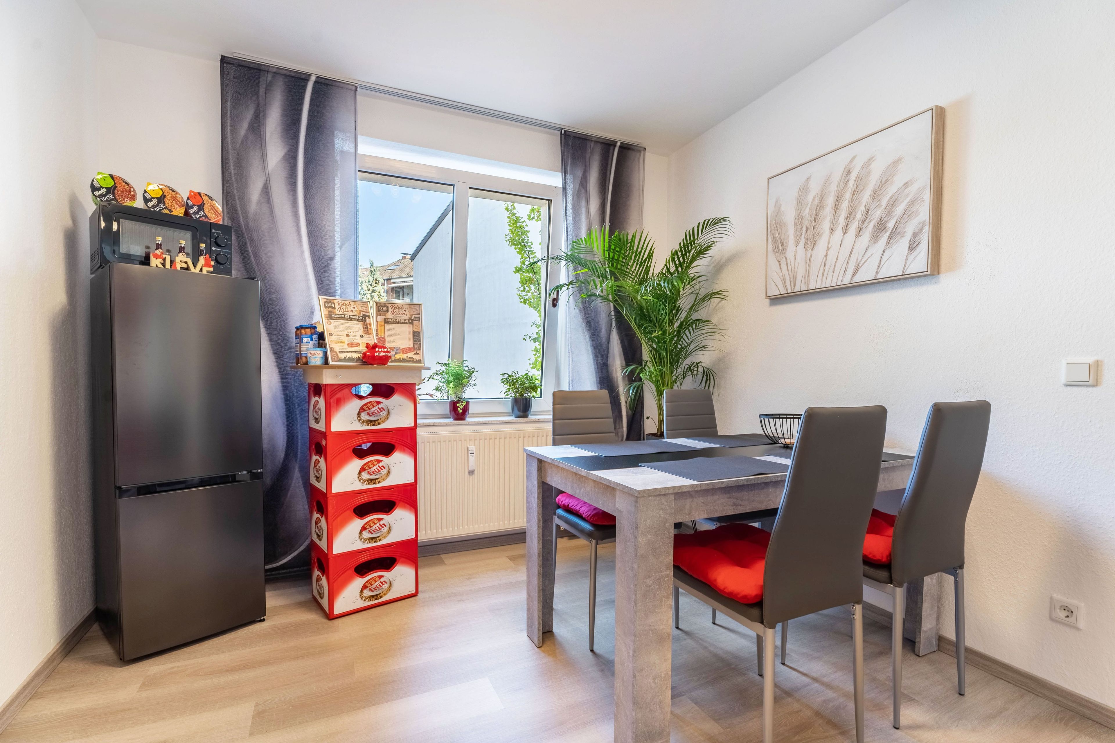 Une cuisine de taille réduite comprenant un réfrigérateur, une table à manger avec des chaises et une fenêtre avec des rideaux.