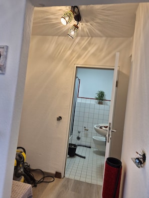 Ein Badezimmer mit gefliestem Boden, einem weißen Waschbecken und einer Duschkabine. Eine Tür führt in einen anderen Raum, und ein Staubsauger steht auf dem Boden.