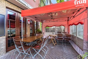 Ulkoterassi, jossa on pöytiä ja tuoleja, tiiliseinä ja punainen markiisi, jossa lukee teksti ’Früh KÖLSCH’.