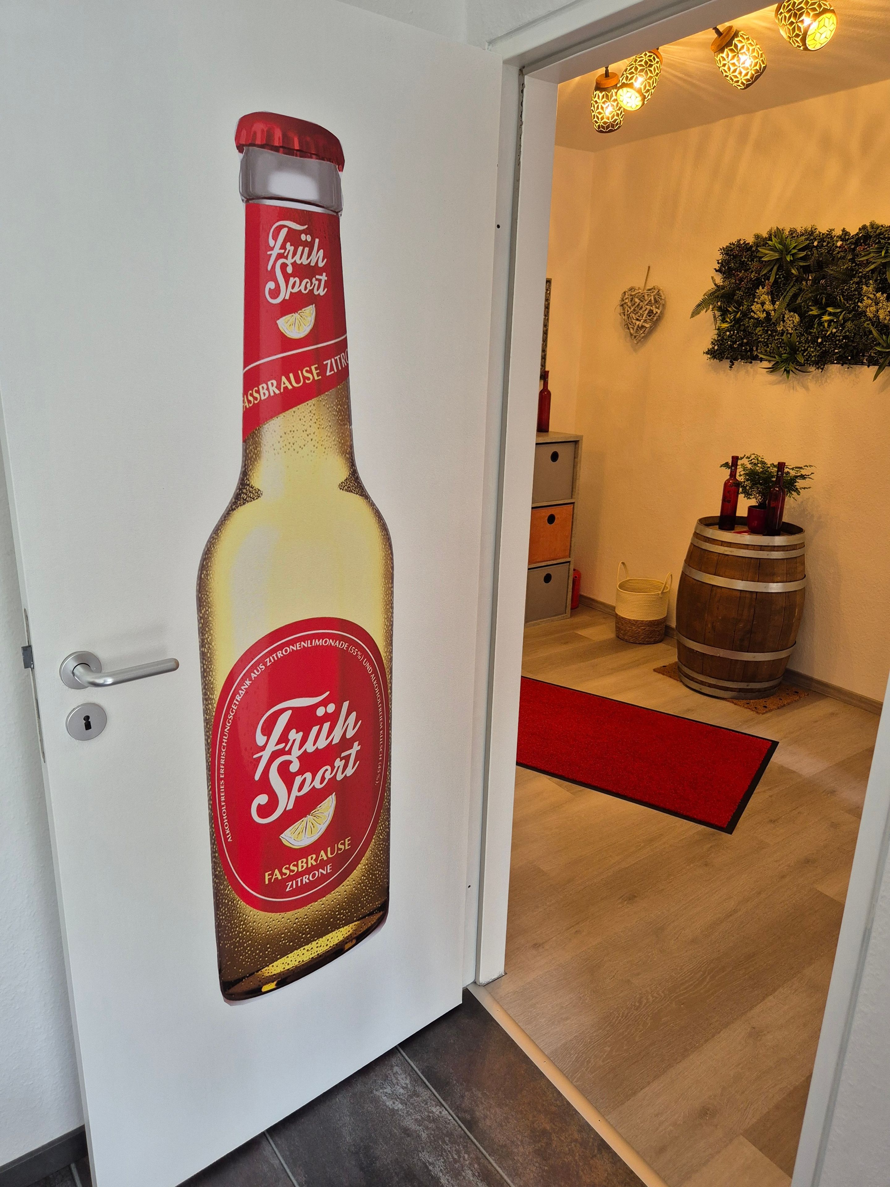 A door with a large sticker of a beer bottle labeled 'Fruh Sport' and a red carpet leading to a room with a wooden barrel and a small plant.