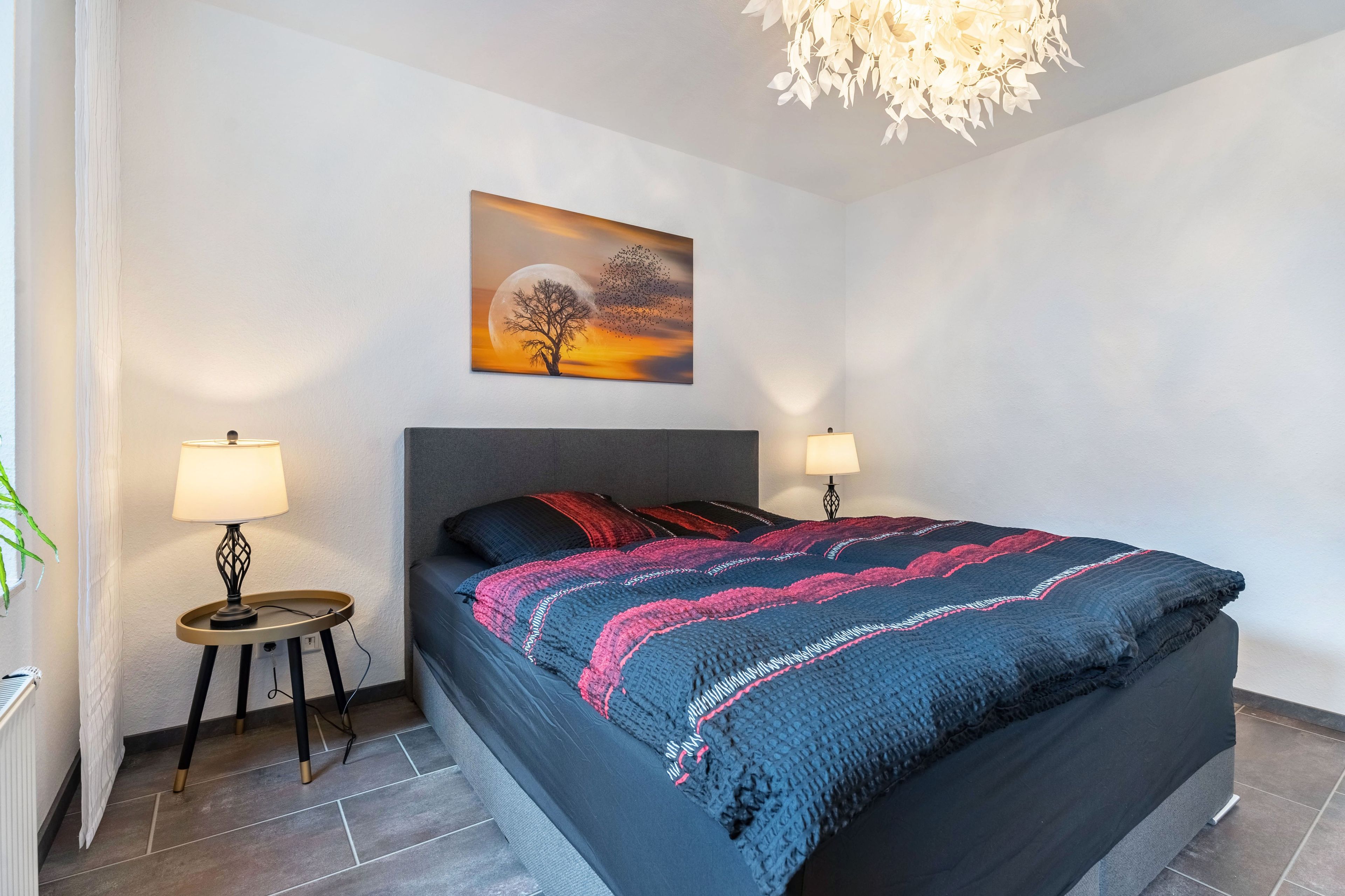 A modern bedroom with a large bed, two bedside lamps, a wall-mounted painting, and a hanging light fixture.