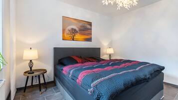 A modern bedroom with a large bed, two bedside lamps, a wall-mounted painting, and a hanging light fixture.