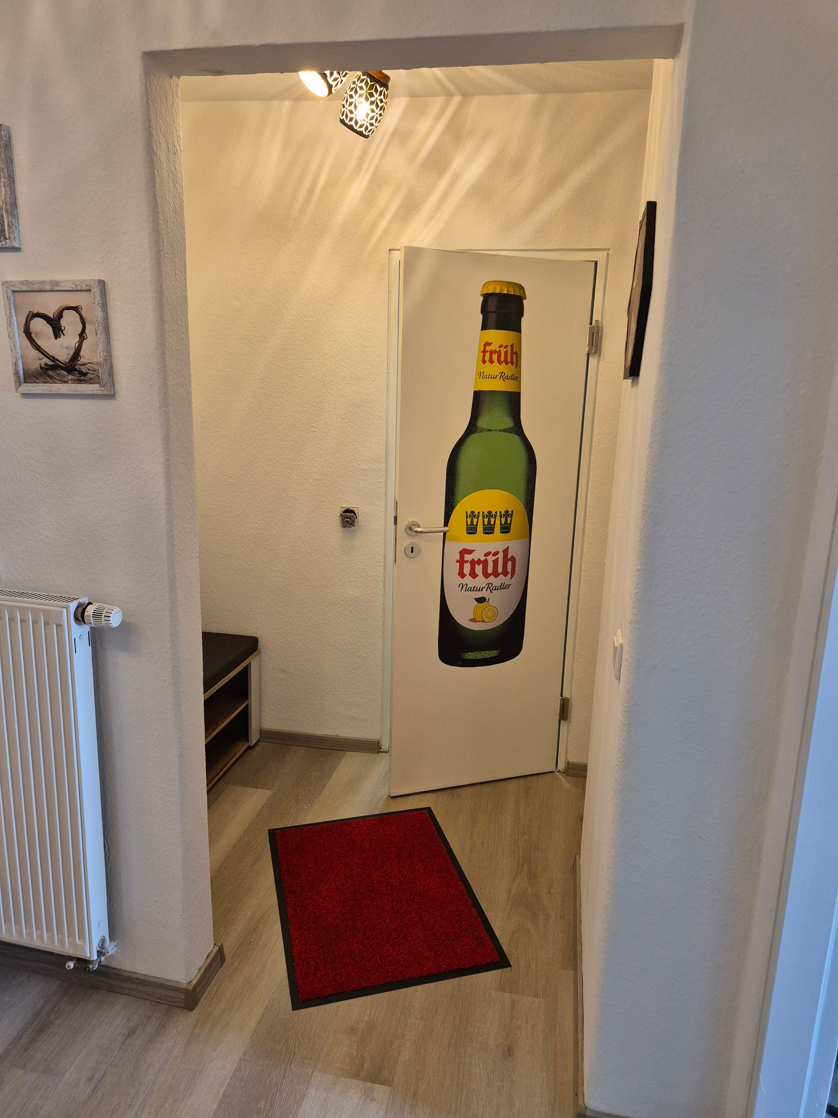 Ein Flur mit einer Tür, die ein Bierflaschen-Design zeigt, einem Heizkörper, einem roten Teppich und einem kleinen Regal.