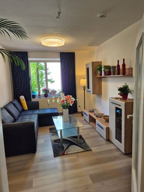 A modern living room with a sofa, a glass coffee table, a kitchen area with a microwave, and a window with curtains.