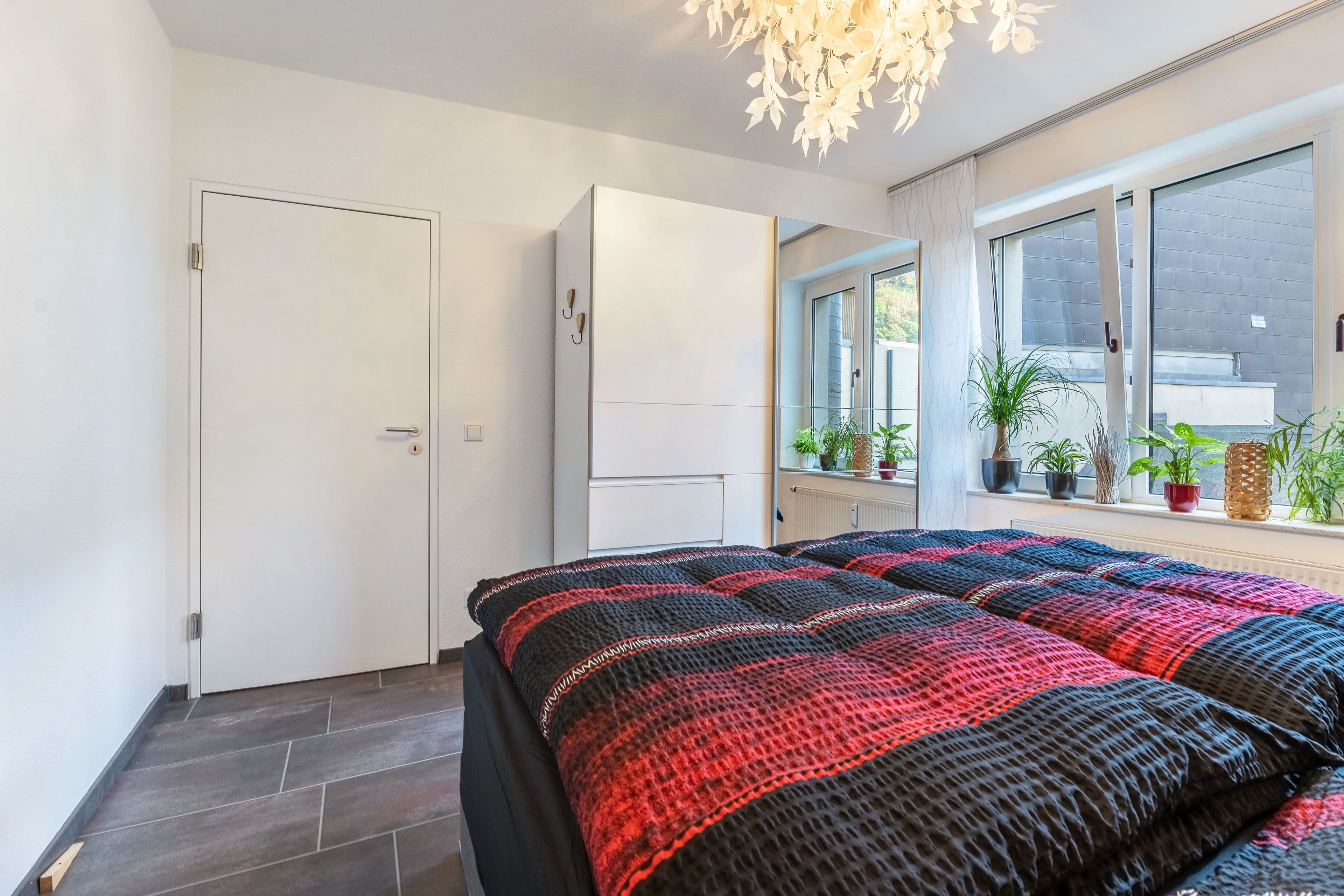 A bedroom with a bed, a wardrobe, a window with plants, and a door.